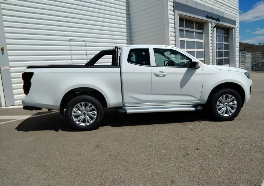 Vue de profil côté droit d'un pick-up Isuzu D-MAX blanc avec arceau noir et couvre-benne roulant, stationné devant un bâtiment industriel.