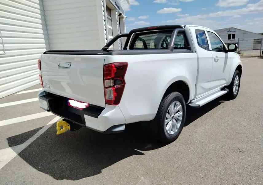 Vue 3/4 arrière droite d’un Isuzu D-MAX blanc avec arceau noir et volet couvre benne roulant, stationné en extérieur.