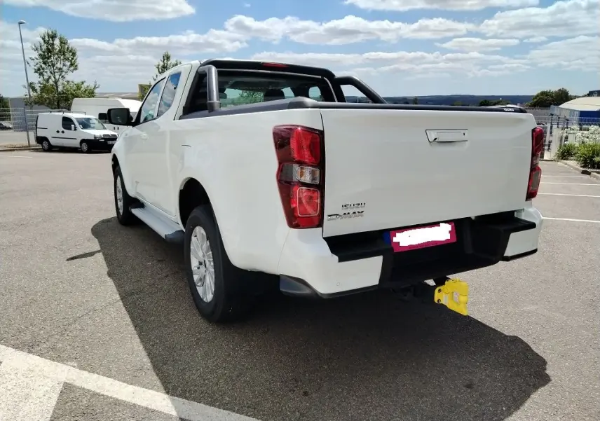 Vue 3/4 arrière droite d'un pick-up Isuzu D-MAX blanc avec arceau et volet couvre benne roulant, attelage jaune visible.