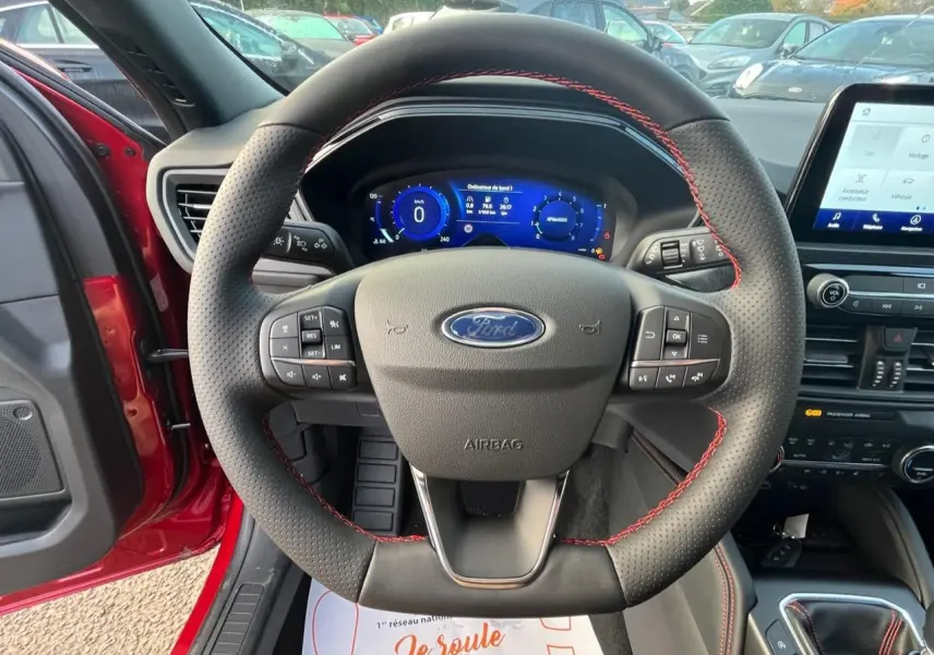 Vue centrée sur le volant perforé avec surpiqûres rouges du Ford Kuga rouge Lucifer, tableau de bord numérique et écran tactile visibles.
