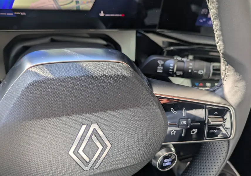 Gros plan sur le volant noir texturé de la Renault Clio VI TCe 115ch Techno, avec commandes intégrées et logo central argenté.