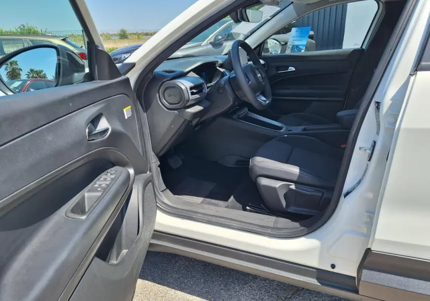 Intérieur côté conducteur du Jeep Avenger blanc 2025, avec tableau de bord et sièges noirs visibles depuis la portière ouverte.