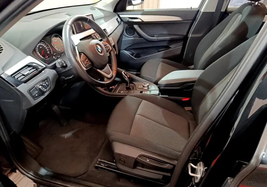 Vue intérieure côté conducteur du BMW X1 noir, montrant le volant cuir multifonctions et la console centrale avec molette.