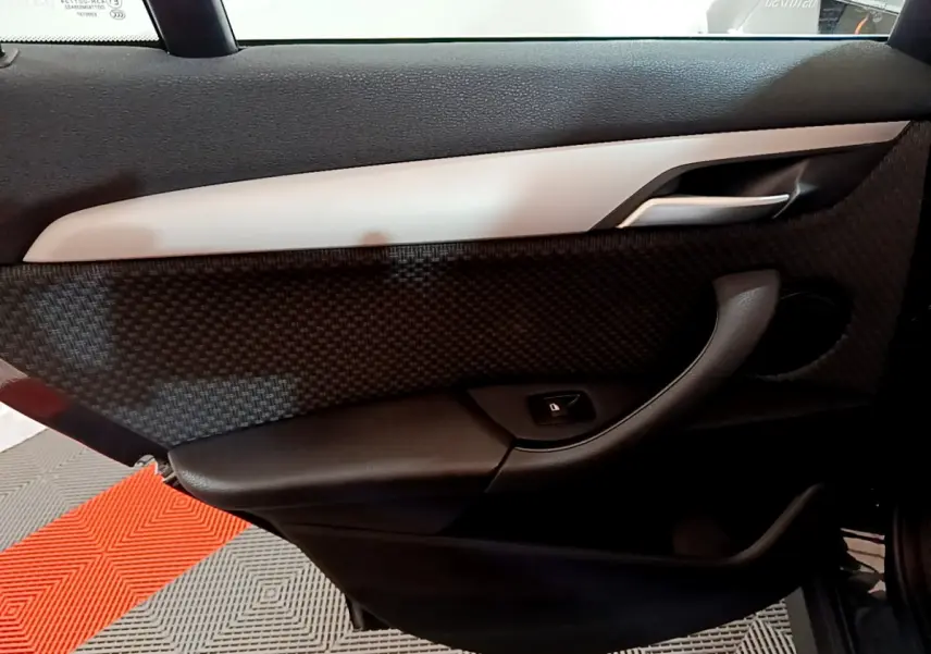Vue intérieure côté gauche de la porte d’une BMW X1 noire, avec garniture noire texturée et poignée argentée.