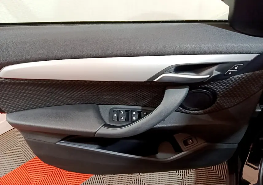 Vue intérieure côté gauche de la porte avant d'une BMW X1 noire, avec garniture carbone et commandes de vitres.