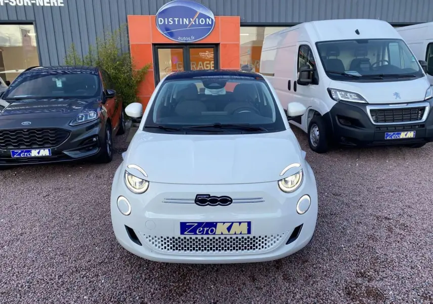 Vue avant d'une Fiat 500 électrique blanche avec phares allumés, stationnée devant un garage Distinxion.
