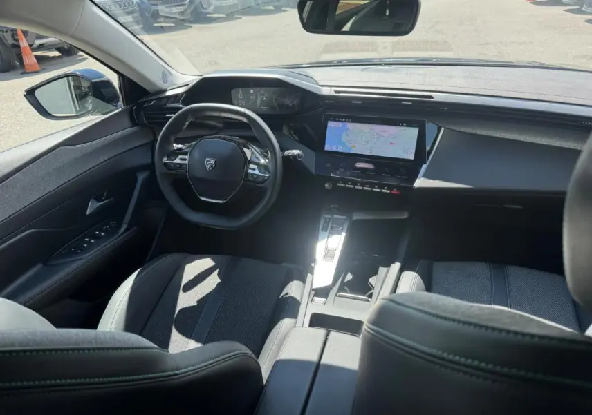 Vue intérieure avant de la Peugeot 308 Hybrid noire, montrant le volant, le cockpit numérique et l'écran central tactile avec navigation.