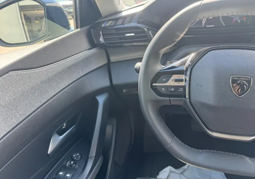 Vue intérieure côté conducteur de la Peugeot 308 Hybrid 2024 noire, volant multifonction et commandes sur porte visibles.