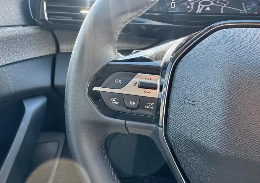 Gros plan sur le volant noir de la Peugeot 308 Hybrid 2024, avec commandes intégrées et cockpit numérique en arrière-plan.