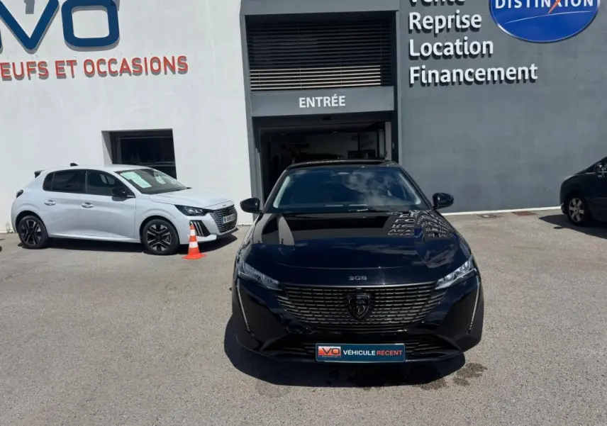 Vue frontale d'une Peugeot 308 Hybrid 136 e-DCS6 Allure noire, garée devant une concession avec reflet du ciel sur la carrosserie.