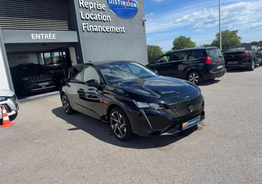 Peugeot 308 Hybrid noir vue 3/4 avant droit, jantes alliage 17 pouces, stationnée devant un garage avec enseigne Distinxion.