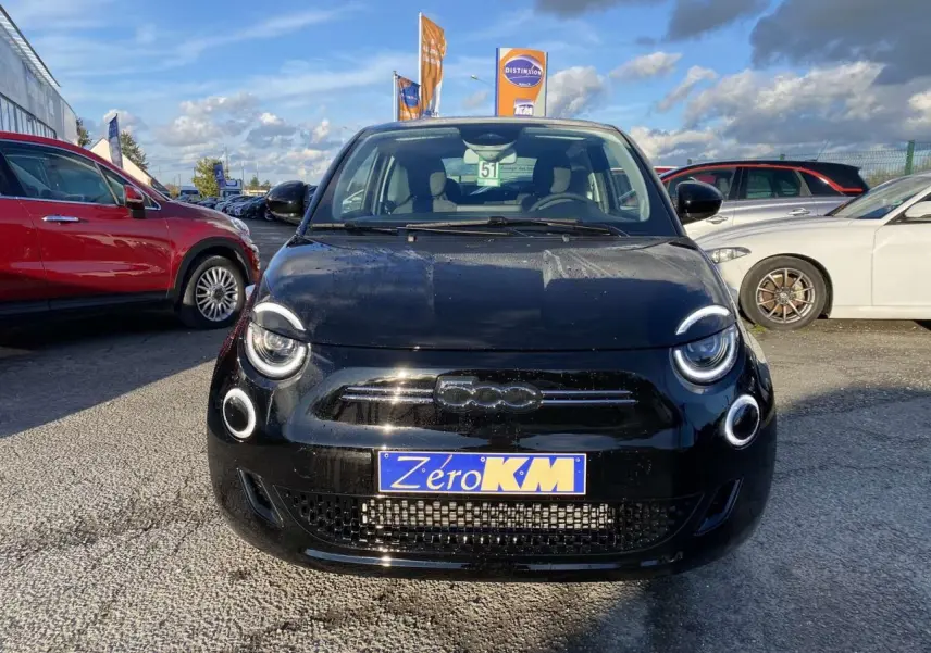 Vue avant d'une FIAT 500 électrique noire avec phares ronds LED et plaque Zéro KM sur un parking extérieur.