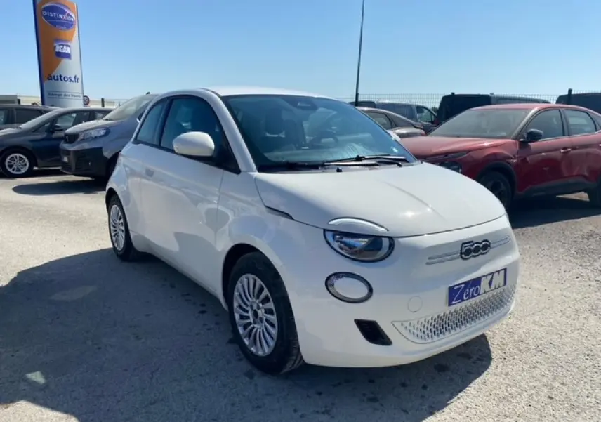 Vue 3/4 avant d'une FIAT 500 électrique blanche avec logo distinctif et plaque "Zero KM" sur un parking extérieur.