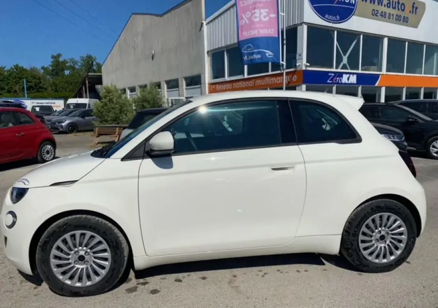 Profil côté gauche d'une FIAT 500 électrique blanche Ice White, avec jantes alliage et toit noir en arrière-plan d'un parking.
