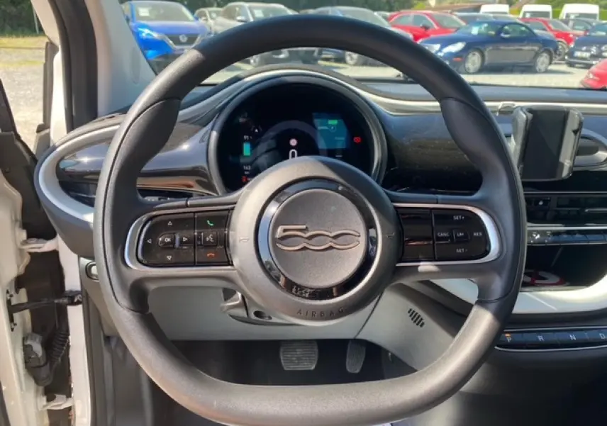 Vue intérieure centrée sur le volant noir de la Fiat 500 électrique blanche avec tableau de bord numérique visible.