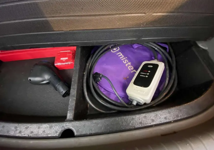 Gros plan sur le compartiment de rangement avant d'une KIA Soul électrique 2016, montrant un câble de recharge et une trousse violette.