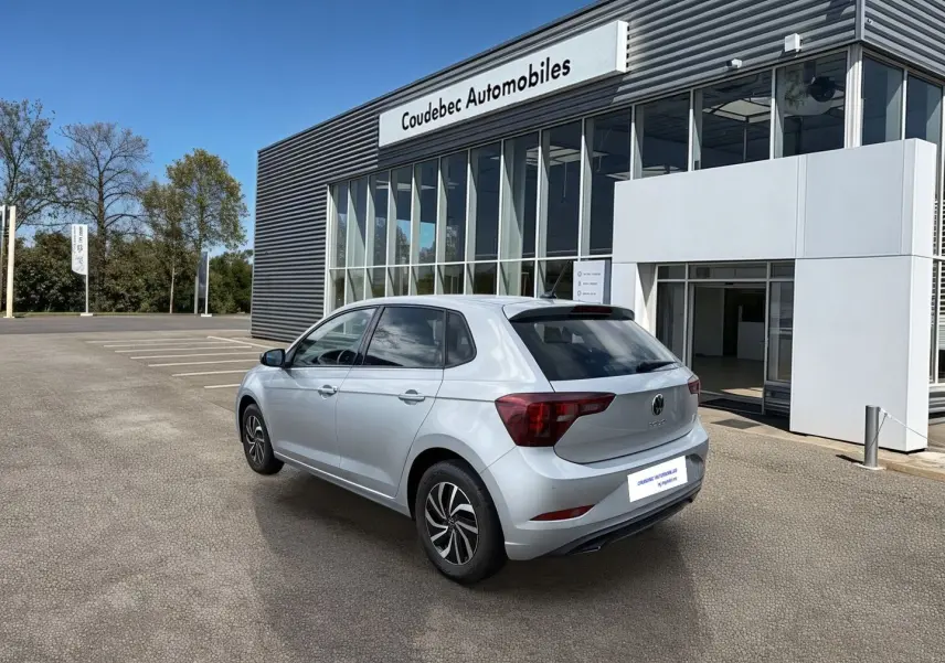 Vue 3/4 arrière droite d'une Volkswagen Polo grise 2025 stationnée devant un bâtiment de concession.