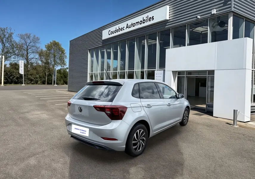 Vue 3/4 arrière droite d'une Volkswagen Polo grise stationnée devant un bâtiment moderne de concession automobile.