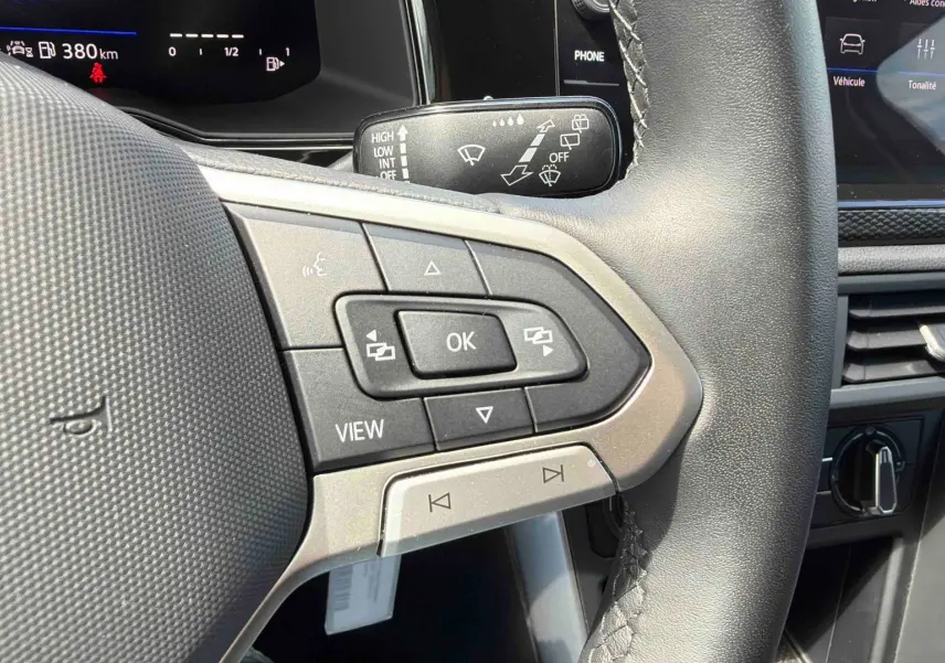 Gros plan sur les commandes au volant noir de la Volkswagen Polo 2025, avec tableau de bord digital en arrière-plan.