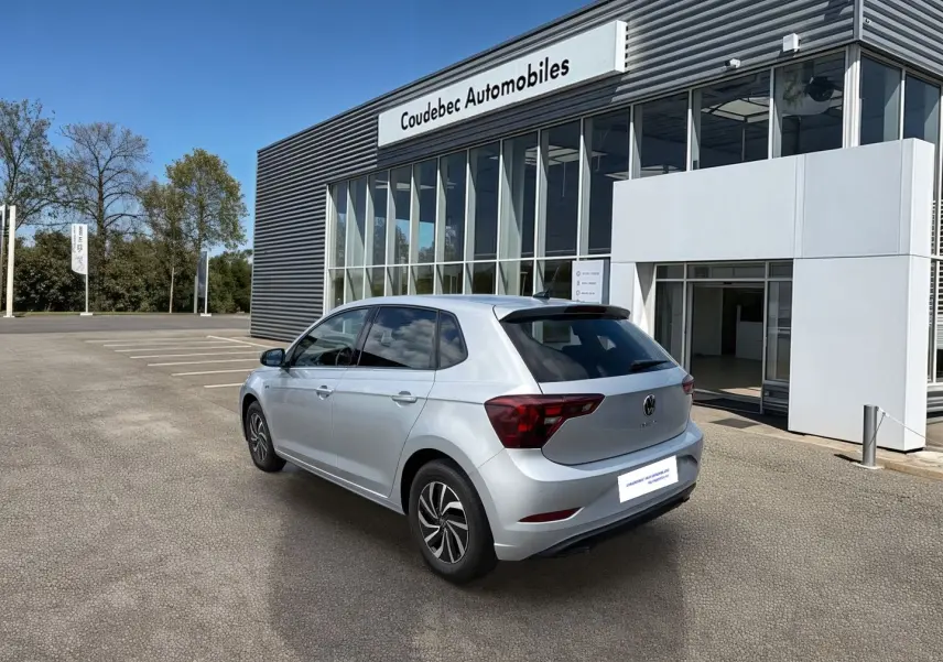 Vue 3/4 arrière droite d'une Volkswagen Polo grise stationnée devant un bâtiment moderne de concession automobile.