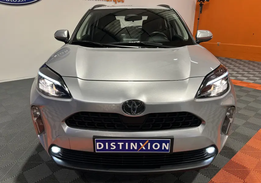 Vue avant d'une Toyota Yaris Cross 116h Active gris minéral avec phares allumés et plaque Distinxion en intérieur showroom.