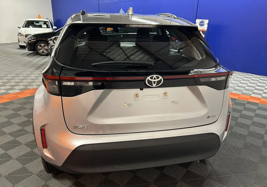 Vue arrière d'une Toyota Yaris Cross 2025 gris minéral avec feux arrière noirs et toit ouvrant en intérieur showroom.