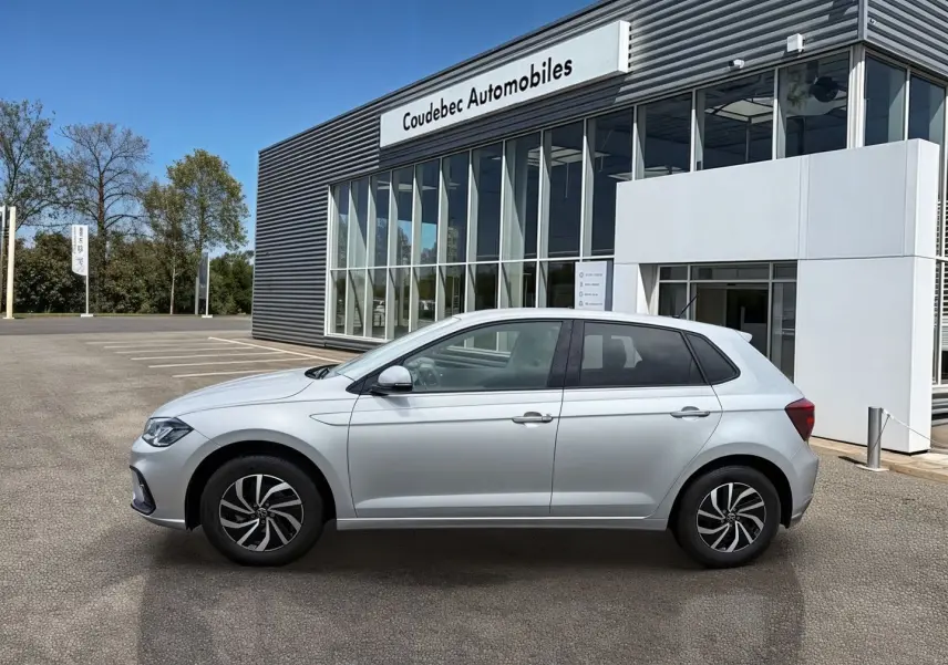 Vue de profil côté gauche d'une Volkswagen Polo grise stationnée devant un bâtiment commercial moderne.