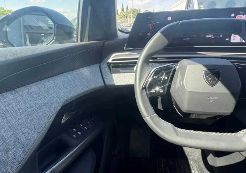 Vue intérieure côté conducteur du Peugeot 3008 Hybrid 2025, volant compact et tableau de bord numérique avec finition tissu gris.