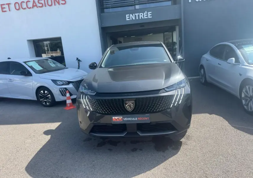 Vue avant d’un Peugeot 3008 Hybrid gris foncé avec calandre noire et feux LED en forme de griffes, stationné devant un bâtiment.