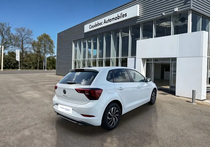 Vue 3/4 arrière droite d’une Volkswagen Polo blanche stationnée devant un bâtiment vitré de concession.