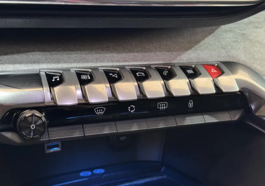 Gros plan sur la rangée de boutons argentés et noirs du tableau de bord du Peugeot 3008 GT gris clair, avec éclairage bleu dessous.