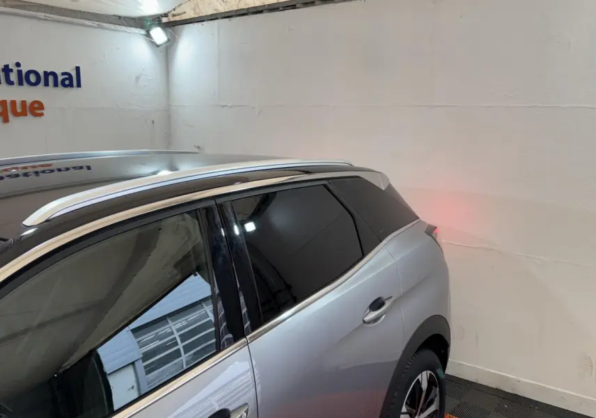 Vue 3/4 arrière droit d’un Peugeot 3008 gris clair avec vitres teintées et barres de toit chromées dans un garage.