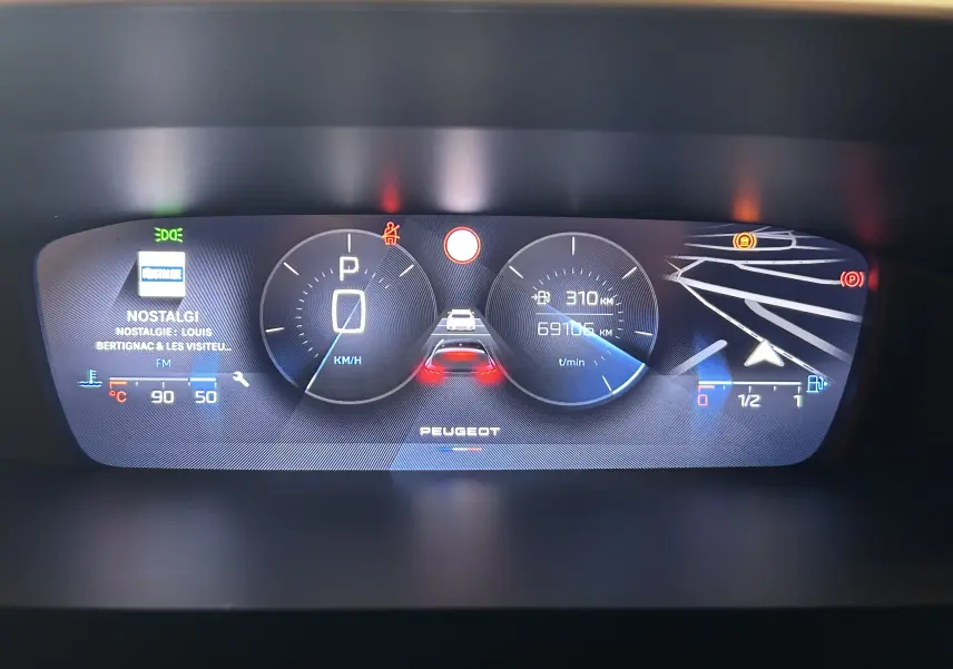 Tableau de bord numérique de Peugeot 308 gris, affichant vitesse, kilométrage, navigation et radio FM Nostalgie.