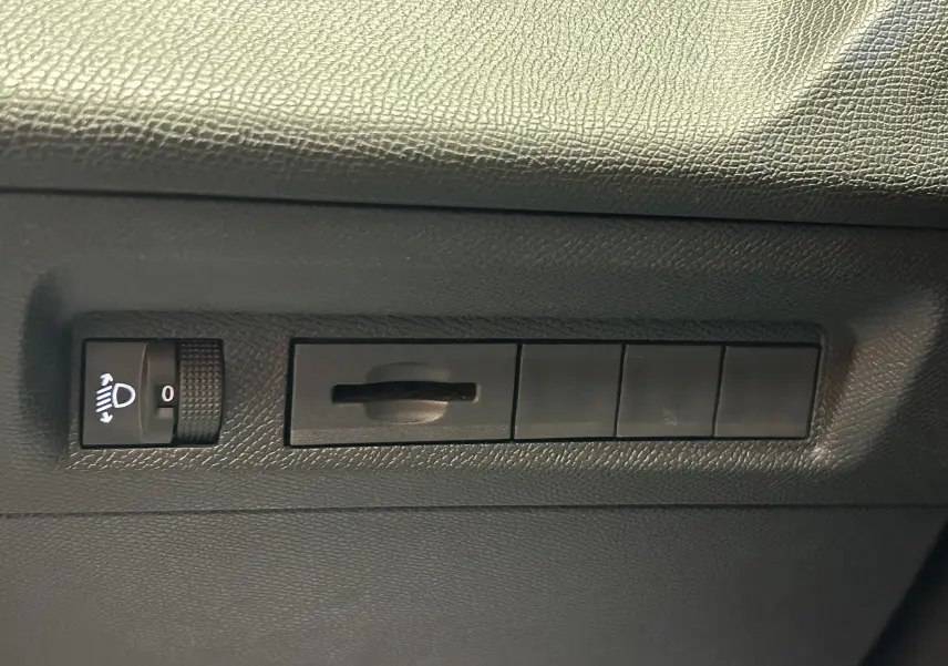 Détail intérieur montrant la molette de réglage des phares et plusieurs boutons noirs sur la planche de bord côté conducteur d’une Peugeot 308 gris.