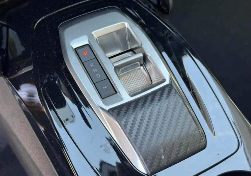 Gros plan sur la console centrale noire brillante de la Peugeot 208 Hybrid 110, avec sélecteur de boîte automatique et finition carbone.