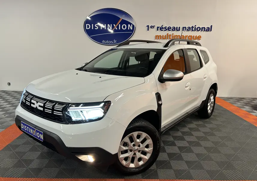 Vue 3/4 avant droite d'un Dacia Duster blanc 2024 avec feux LED allumés et barres de toit noires.