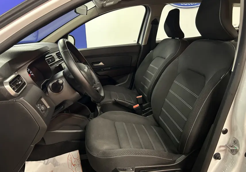 Vue intérieure côté conducteur du Dacia Duster blanc 2024, mettant en valeur les sièges chauffants en tissu noir avec coutures contrastées.