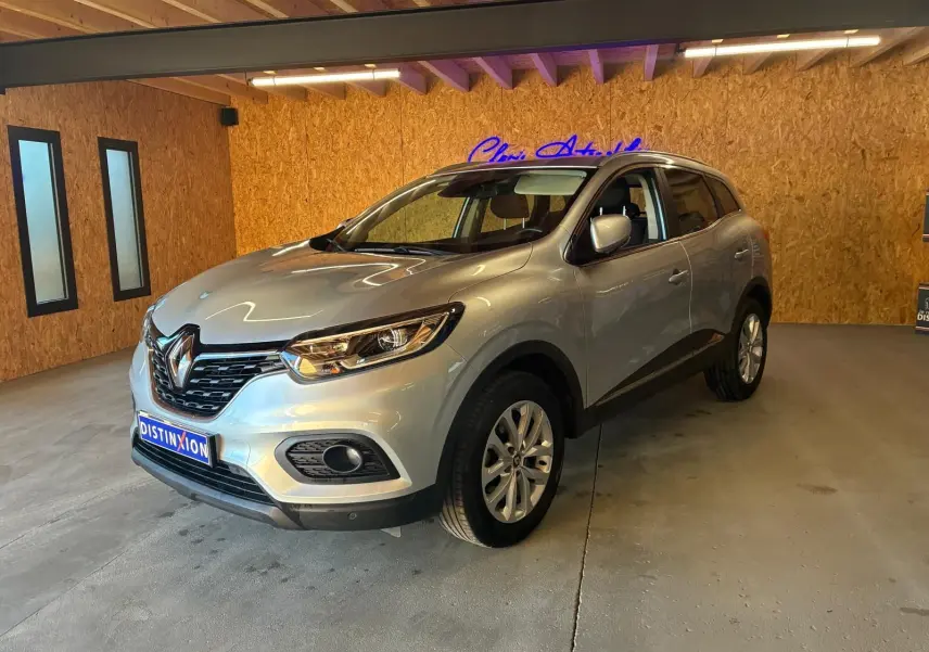 Renault Kadjar gris en 3/4 avant droit dans un garage avec éclairage intérieur et logo néon bleu en arrière-plan.