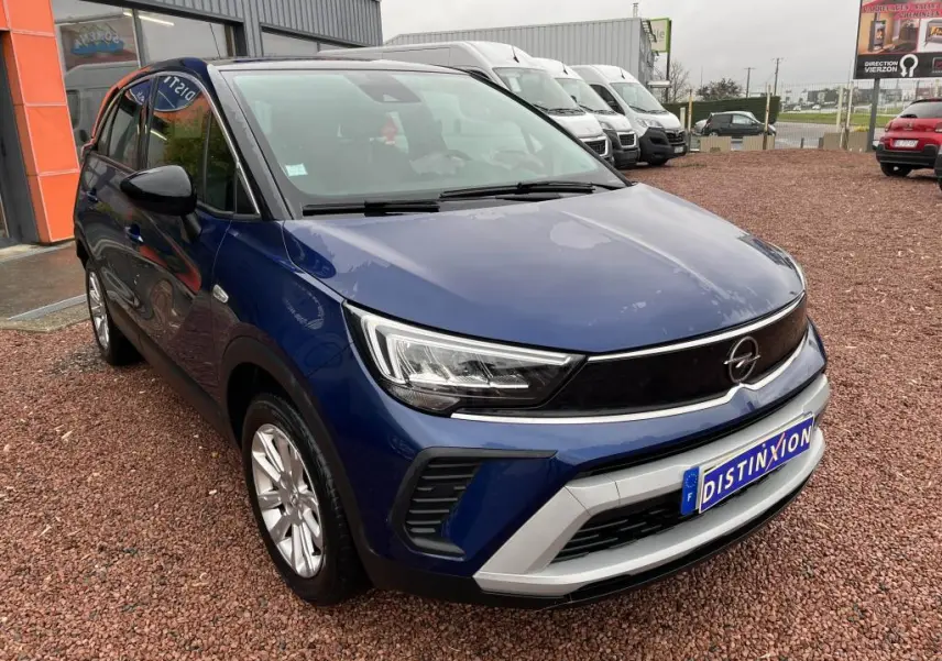 Vue 3/4 avant d'un Opel Crossland bleu clair avec toit noir, garé sur un sol en gravier devant un bâtiment.