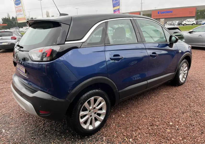 Vue 3/4 arrière droite d'un Opel Crossland bleu clair avec toit noir, garé sur un sol en gravier rouge.