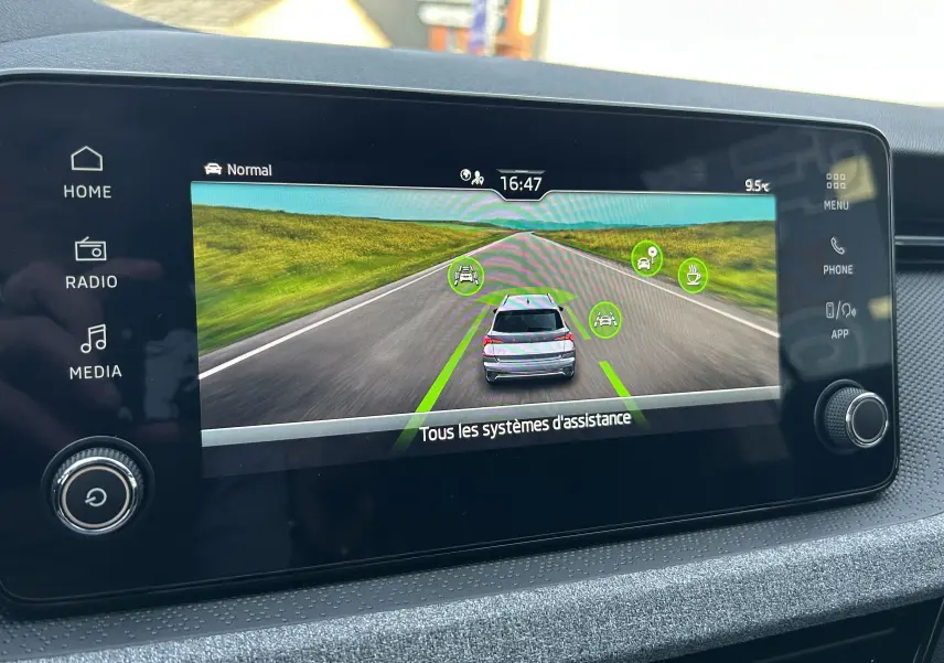 Écran tactile intérieur montrant l'assistance à la conduite avec une Skoda Kamiq gris argent sur route virtuelle.