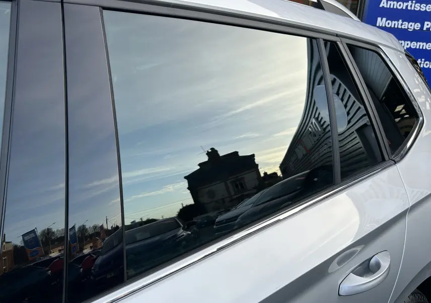 Vue latérale droite sur la porte arrière d’un Skoda Kamiq gris argent métallisé avec vitre teintée reflétant le ciel et un bâtiment.