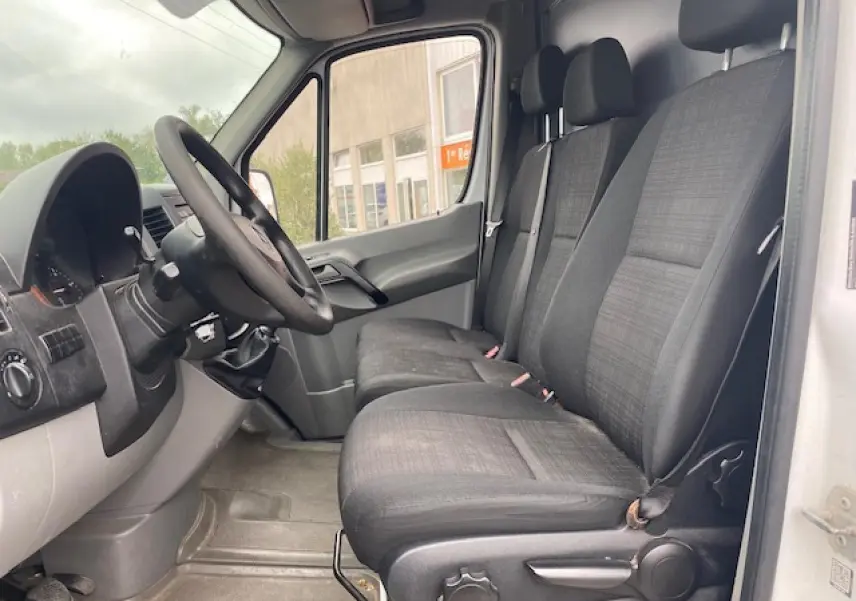 Vue intérieure côté conducteur du Mercedes Sprinter Fourgon blanc de 2015, montrant les sièges avant et le volant noir.