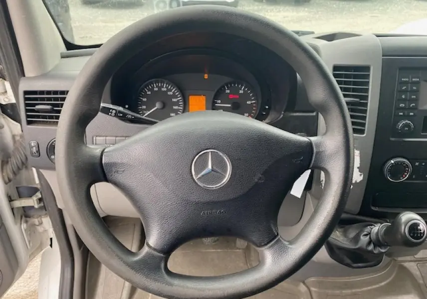 Vue intérieure centrée sur le volant noir Mercedes d'un Sprinter Fourgon blanc avec tableau de bord et levier manuel.