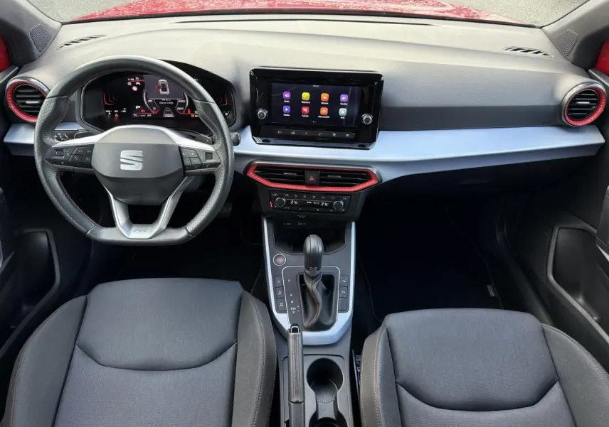 Vue intérieure avant du tableau de bord du SEAT Arona rouge, avec volant sport cuir perforé et écran tactile central.