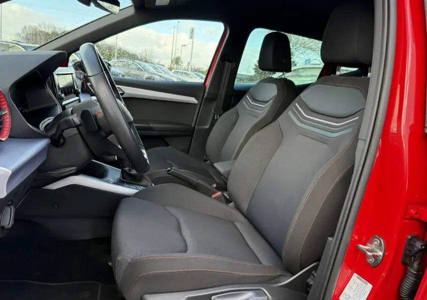 Vue intérieure côté conducteur du SEAT Arona rouge 2024, mettant en valeur les sièges avant noirs avec surpiqûres rouges et volant sport.