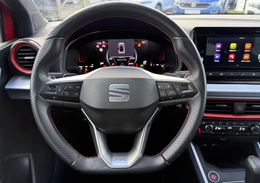 Vue rapprochée du volant sport en cuir perforé noir avec surpiqûres rouges et tableau de bord digital d'une SEAT Arona rouge 2024.