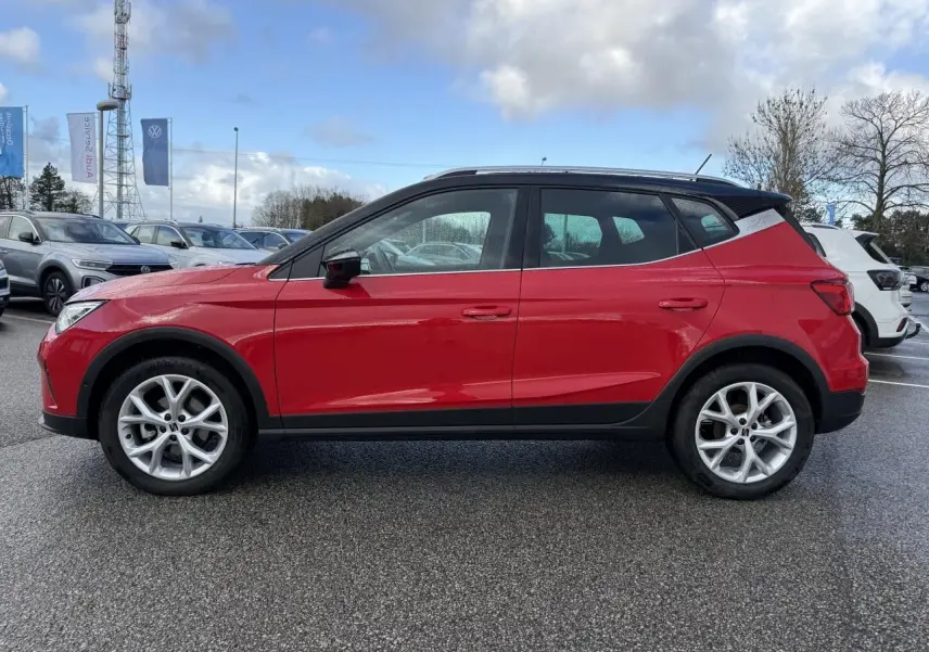 Vue de profil côté gauche d’un SEAT Arona rouge 2024 avec toit noir et jantes argentées sur parking.
