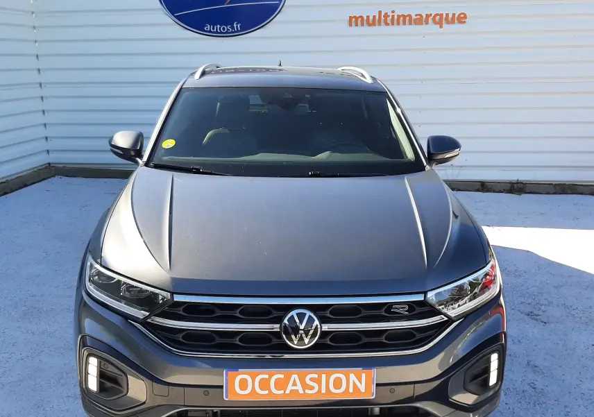 Vue frontale d'un Volkswagen T-Roc gris Indium 2024 avec calandre R-Line et feux LED allumés, sur fond de panneau Distinxion.