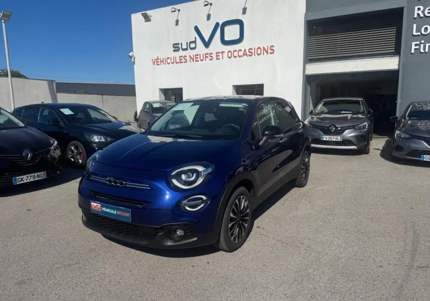 Fiat 500X bleu en 3/4 avant droit, avec jantes classiques et phares ronds distinctifs, stationnée devant un concessionnaire.